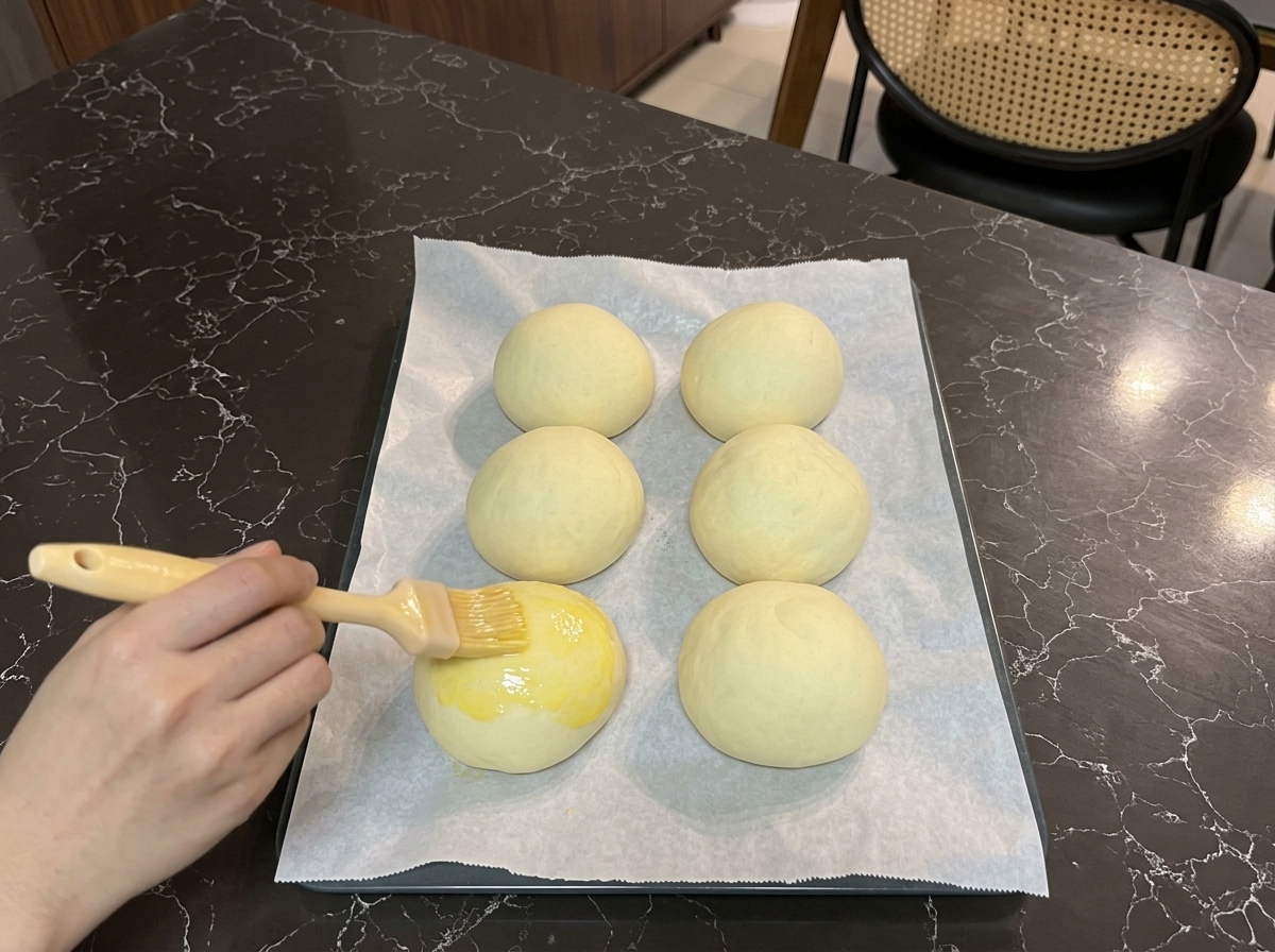 Une main passant un pinceau à pâtisserie sur le dessus dun pain à burger levé avec une dorure brillante à lœuf.