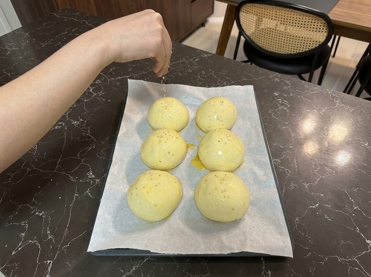 Une main saupoudrant de petites graines de sésame blanc sur les pâtons dorés disposés sur la plaque.