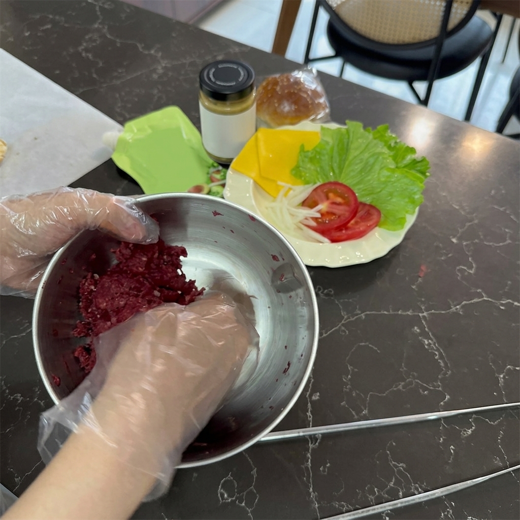Des mains gantées de plastique transparent mélangeant du bœuf haché cru et bien rouge dans un grand bol en inox.