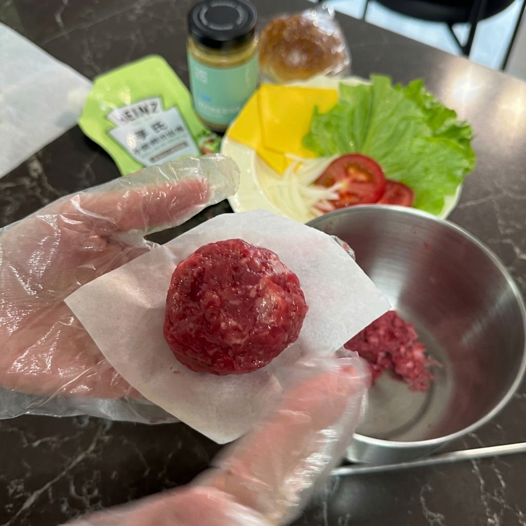 Une main gantée tenant une boulette de bœuf haché cru posée sur un petit carré de papier sulfurisé blanc.