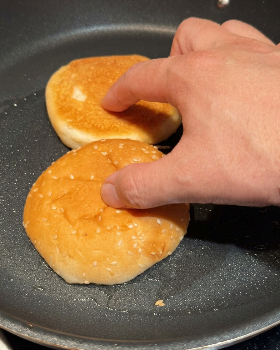 Une main pressant légèrement les deux moitiés dun pain à burger dans une poêle chaude pour les faire dorer.