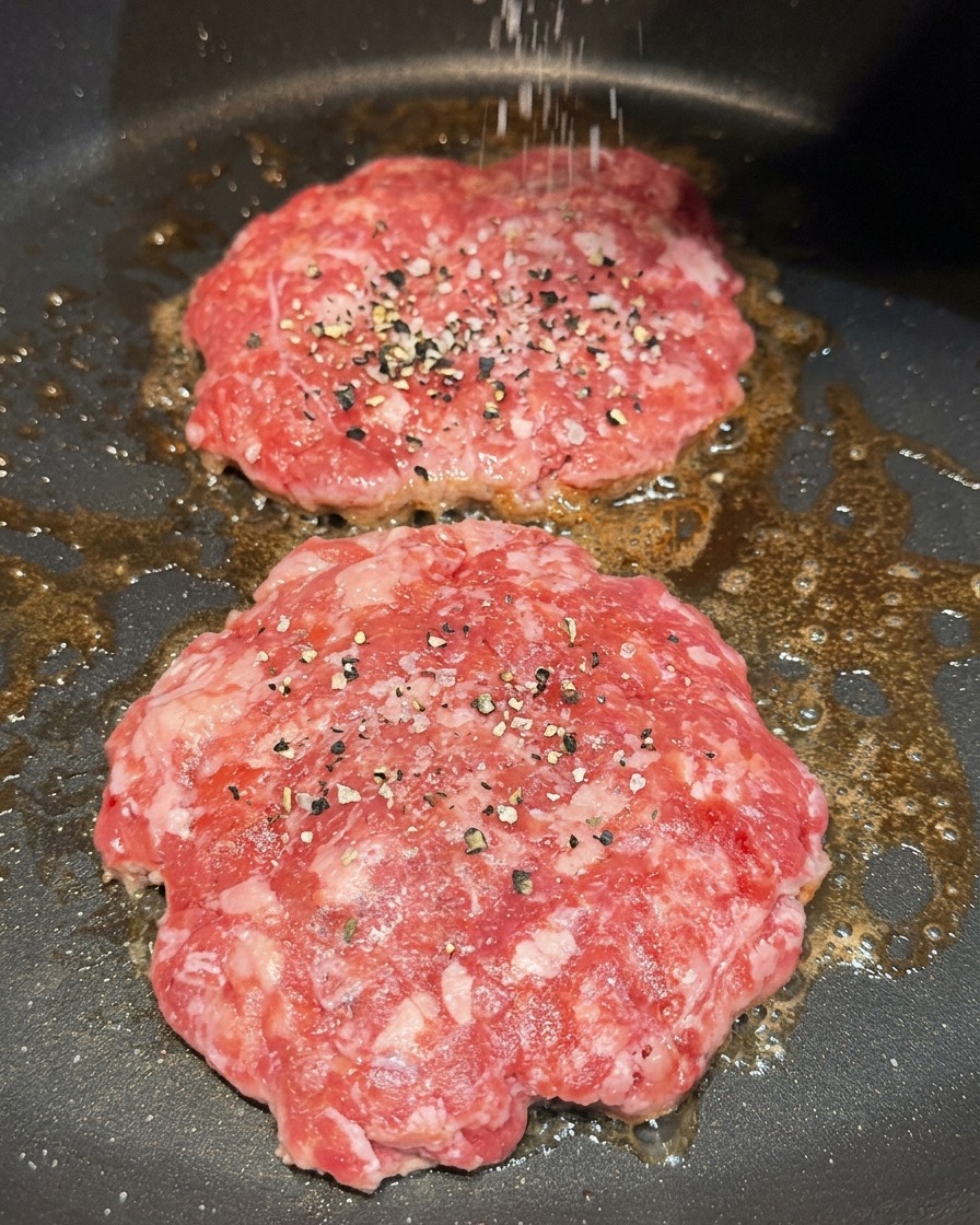 Du gros sel et du poivre noir moulu saupoudrés sur deux steaks hachés en train de cuire dans une poêle.