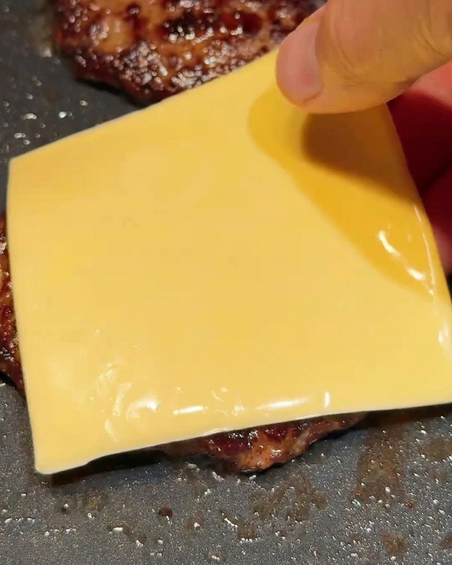 Une main déposant une tranche carrée de fromage sur un steak haché bien chaud dans une poêle.