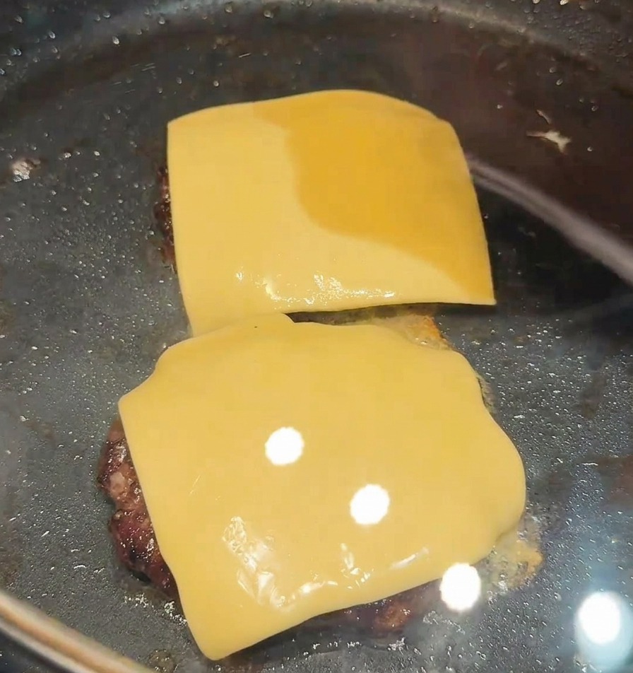 Deux tranches de fromage fondues sur deux steaks rissolant dans une poêle.