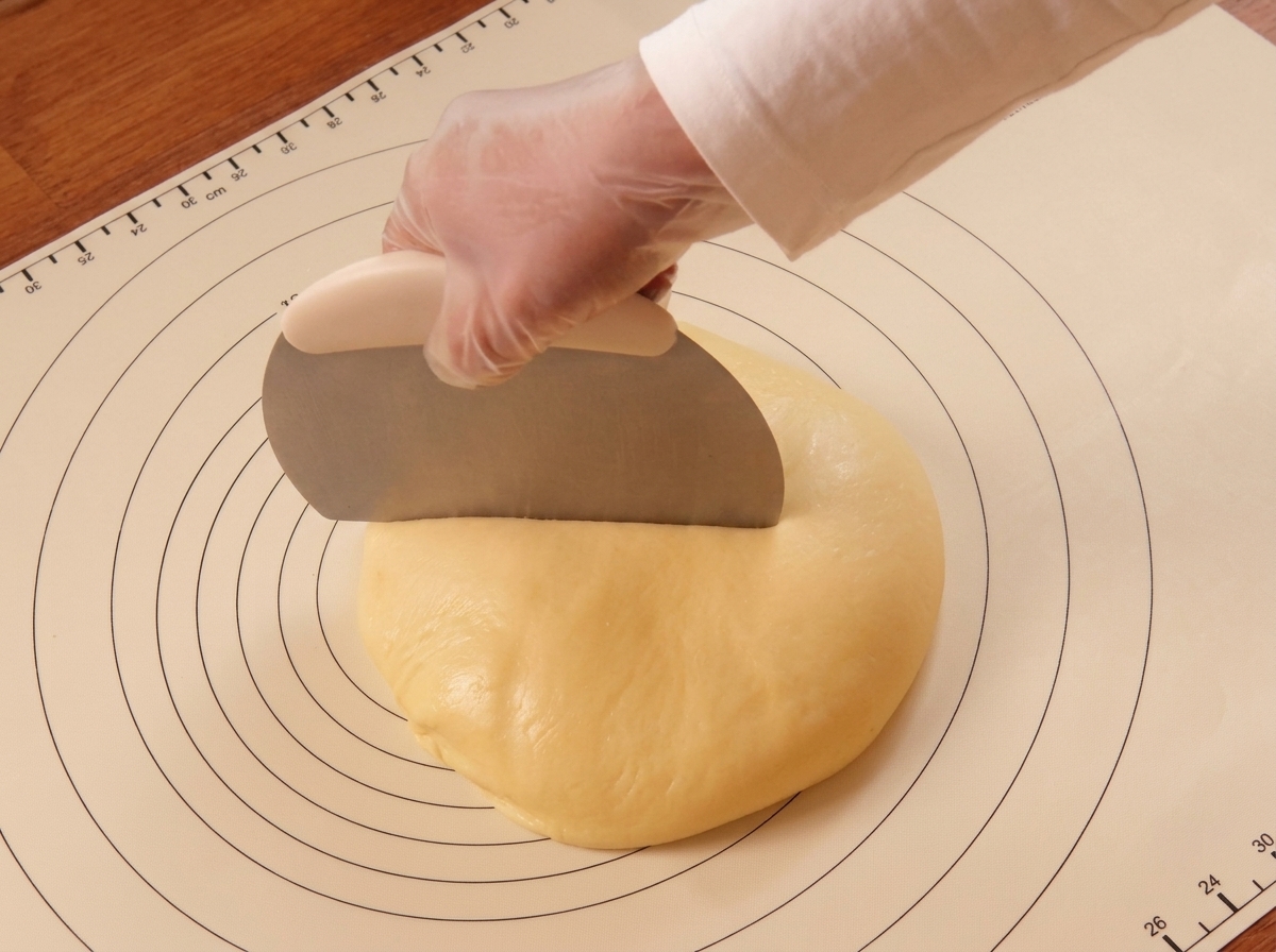Une main gantée utilisant un coupe-pâte en métal pour diviser un pâton sur un tapis de cuisson.