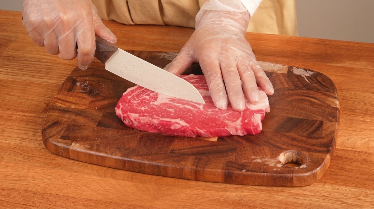 Des mains gantées utilisant un couteau pour trancher une pièce de bœuf persillée sur une planche en bois.