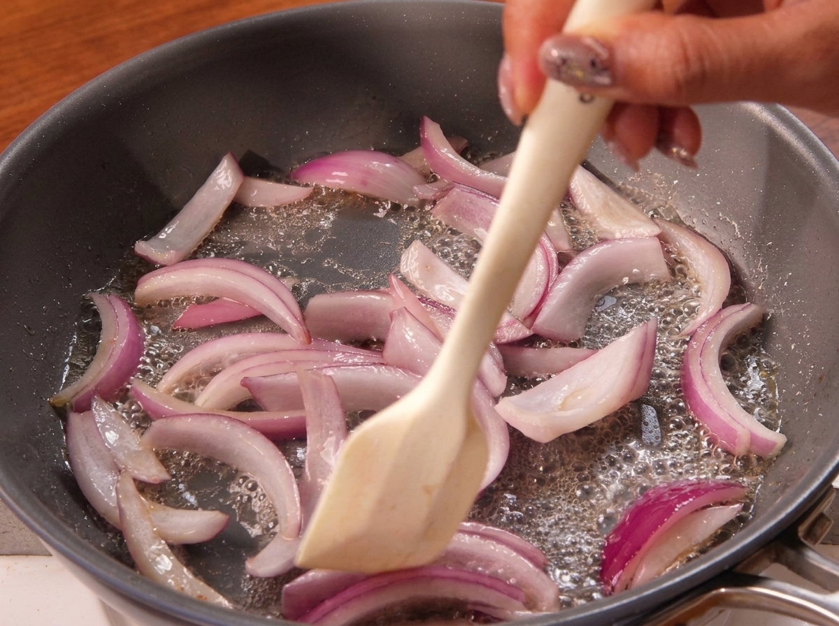Une spatule remuant des oignons rouges émincés qui dorent dans une poêle.