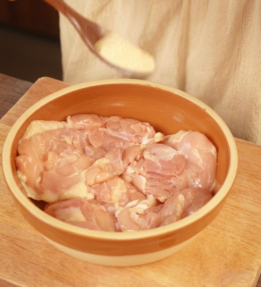 Une cuillère en bois saupoudrant de lail en poudre sur des hauts de cuisses de poulet crus dans un bol en céramique.