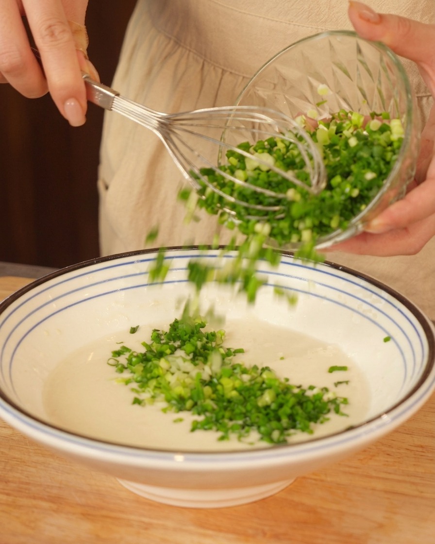 Verser des oignons verts hachés dans une pâte blanche tout en tenant un fouet.
