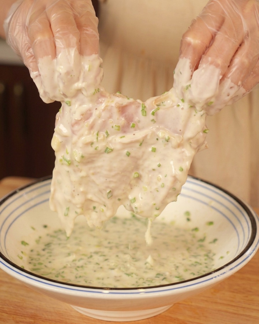 Mains gantées soulevant un haut de cuisse de poulet recouvert dune pâte épaisse parsemée de vert.