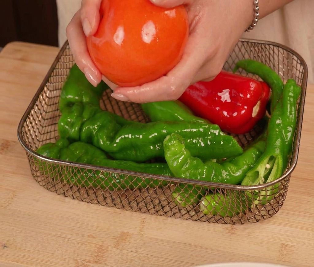 Mains plaçant une tomate huilée et plusieurs piments verts et rouges dans le panier métallique dune friteuse à air.