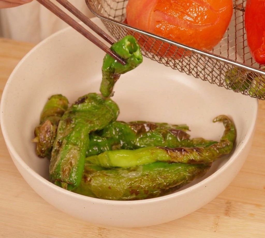 Baguettes soulevant un piment vert grillé du panier dune friteuse à air contenant une tomate rôtie et dautres piments.
