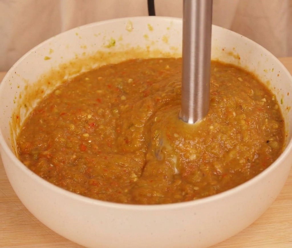 Un mixeur plongeant transformant les piments et la tomate en une sauce épaisse et texturée de couleur orangée.