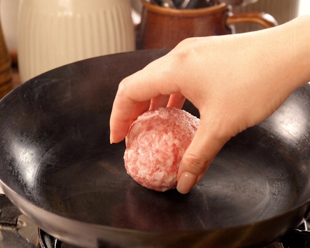 Une main déposant une boule de bœuf haché dans un wok en fonte fumant et non graissé.