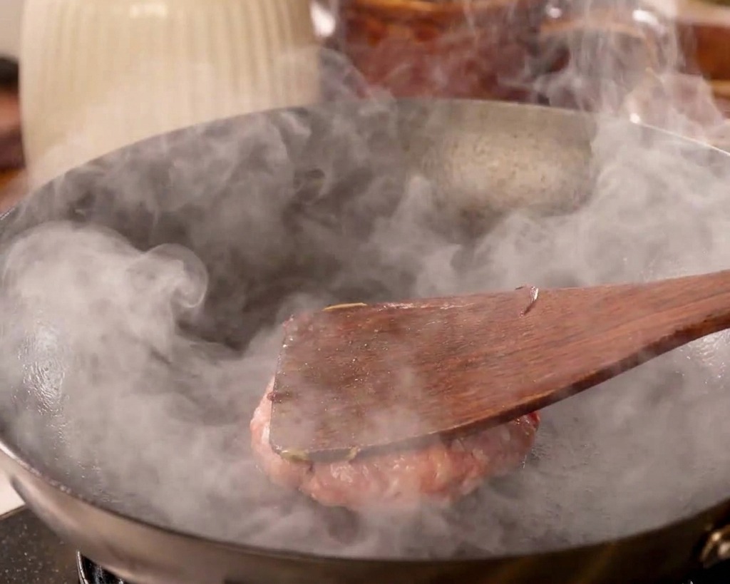 Une spatule plate en bois pressant fermement une boule de bœuf pour laplatir contre la paroi dun wok fumant.