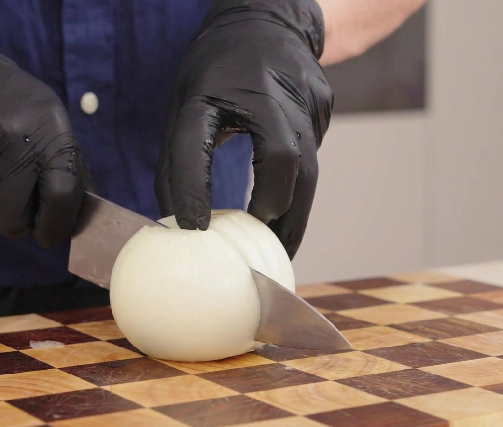 Mains gantées tenant un couteau pour couper un oignon blanc en lanières sur une planche en bois.