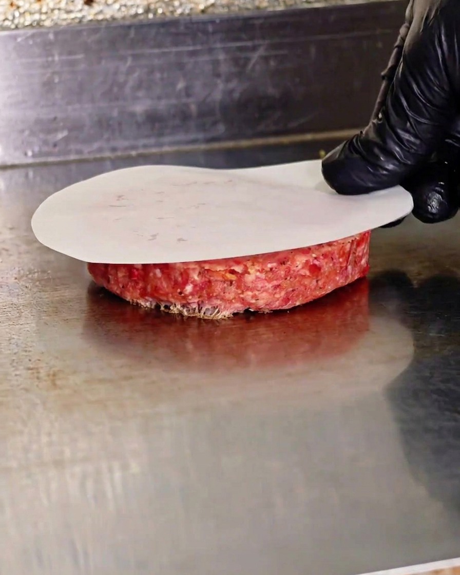 Une main pressant un steak de bœuf cru à laide dune presse plate sur du papier sulfurisé sur une plaque chaude.