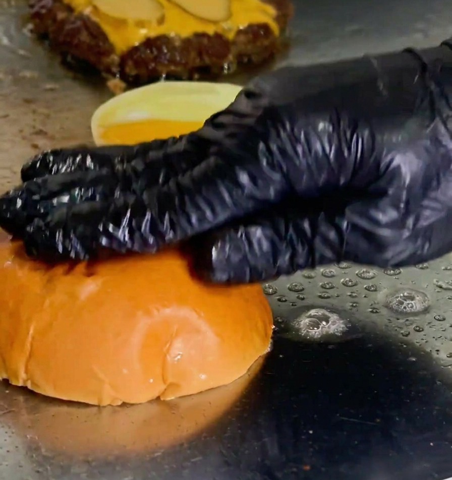 Une main pressant un pain à burger face contre plaque dans du beurre fondu à côté dun steak en cuisson.