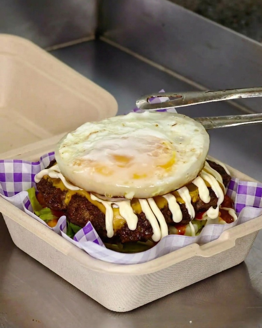 Des pinces déposant un œuf au plat sur un burger garni de viande, fromage et mayonnaise.