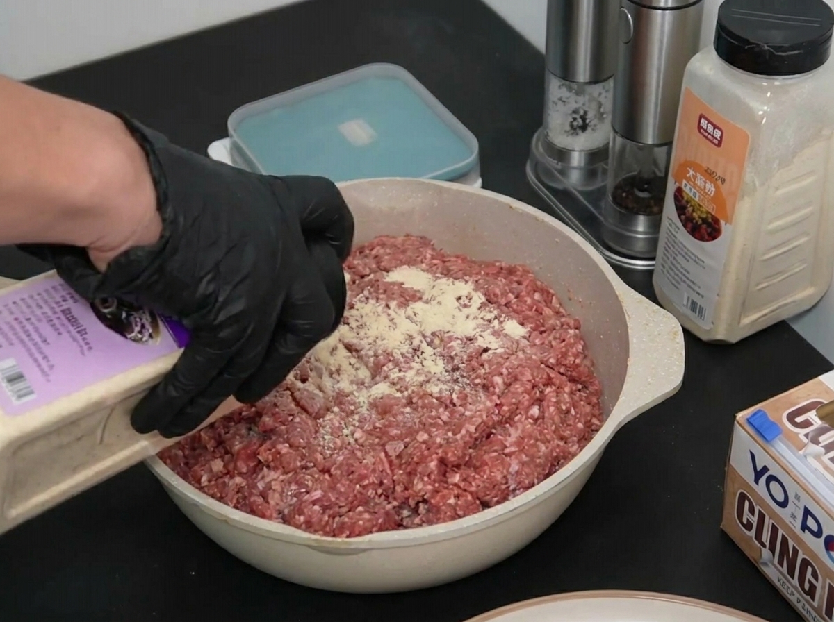 Une main gantée saupoudrant de loignon en poudre sur de la viande de bœuf hachée crue dans un plat.