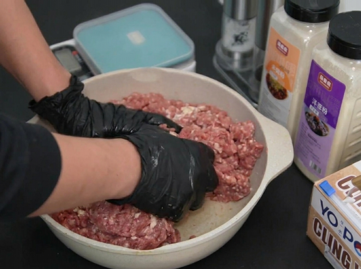 Deux mains gantées malaxant et mélangeant le bœuf haché dans un plat.