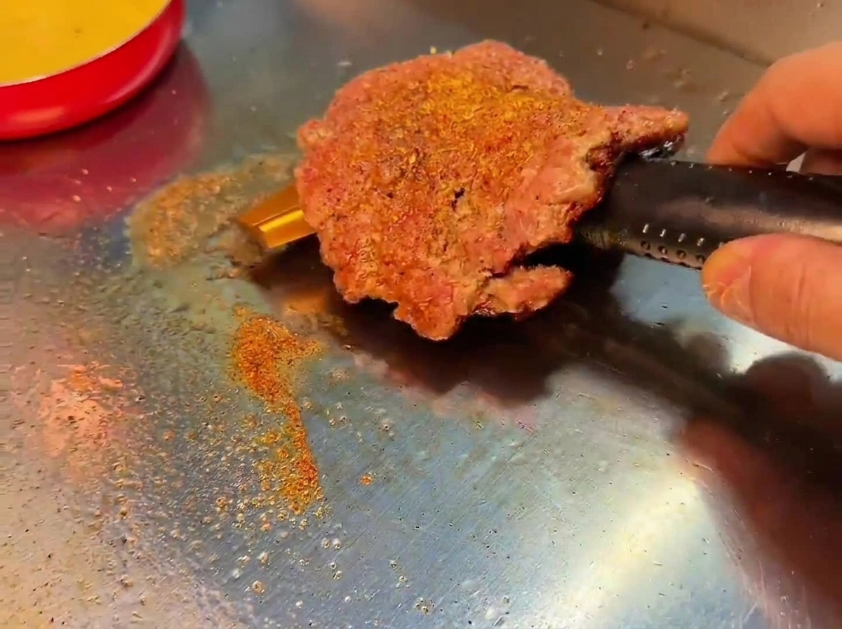 Un steak de bœuf smashé retourné sur le grill, révélant une croûte bien brune et assaisonnée.