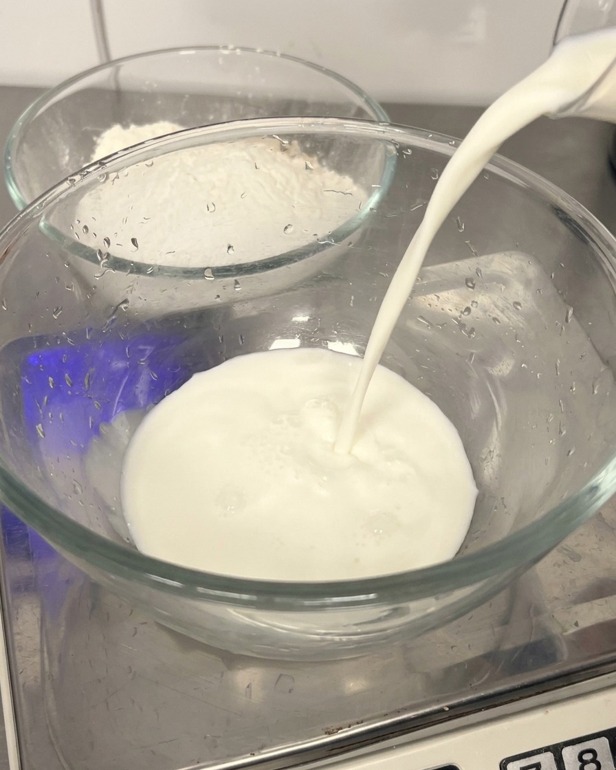 Lait frais versé dans un bol en verre transparent avec un bol de farine en arrière-plan.