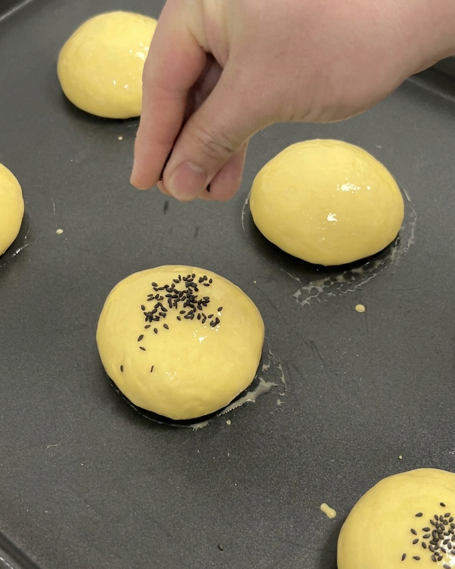 Une main saupoudrant des graines de sésame noir au centre dun pain brioché doré sur une plaque de cuisson.