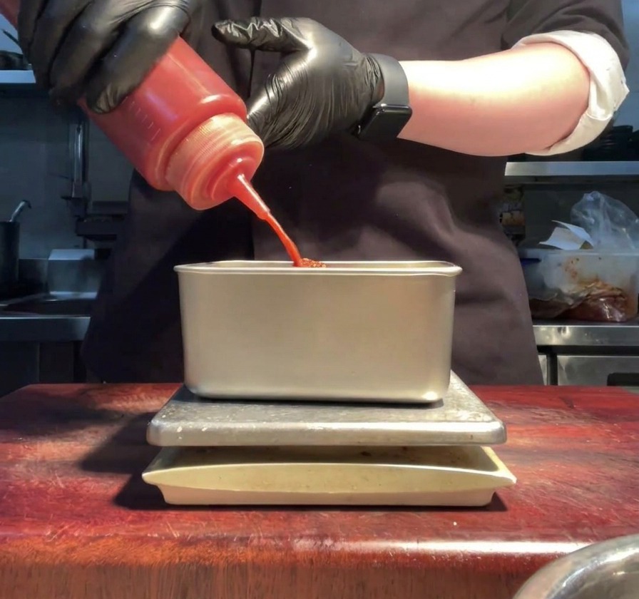 Mains gantées versant de la sauce sriracha rouge vif dune bouteille dans un récipient de préparation en métal.