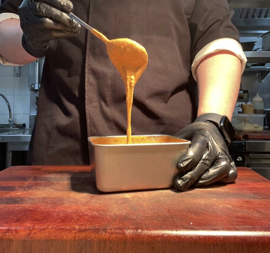 Une main gantée soulevant une cuillerée de sauce burger orange vif et onctueuse.
