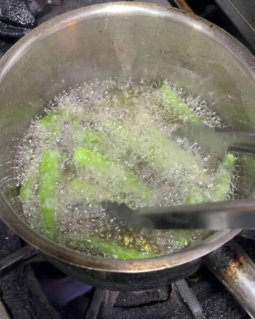 Pinces métalliques manipulant des piments verts entiers en train de frire dans une casserole dhuile chaude.