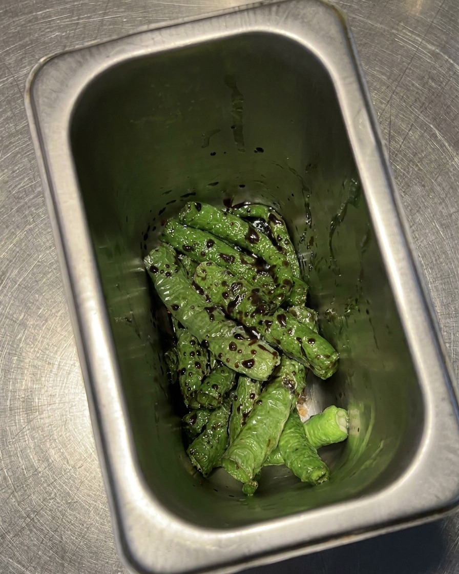 Piments verts grillés dans un récipient en métal, enrobés dun mélange sombre de vinaigre et de sauce soja.