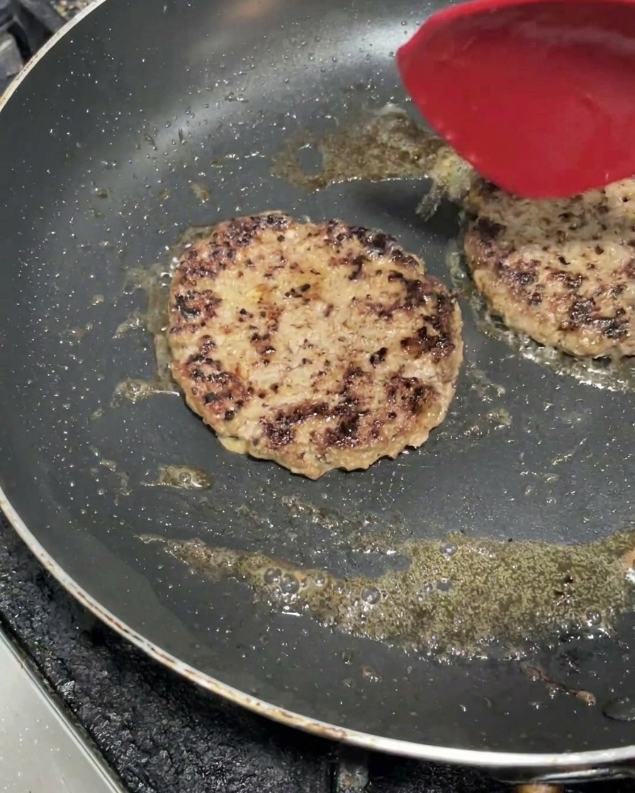Deux steaks de bœuf épais grésillant dans une poêle chaude, développant une croûte brune avec une spatule rouge.