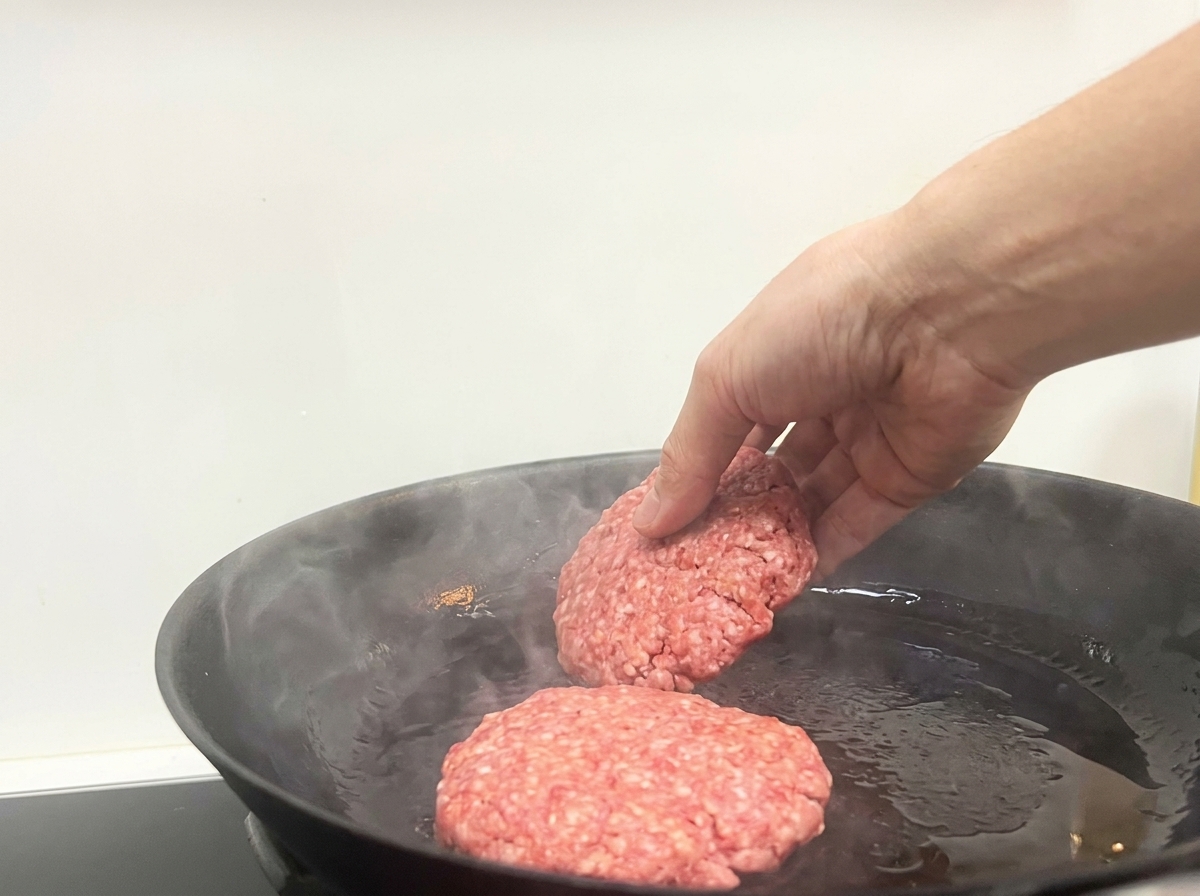 Une main déposant deux steaks de bœuf crus dans une poêle noire fumante.