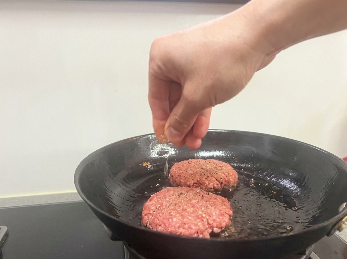 Une main saupoudrant du sel et du poivre noir sur deux steaks de bœuf saisissant dans une poêle.