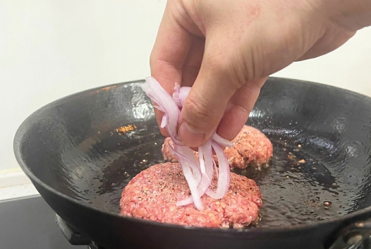 Une main déposant des oignons rouges émincés sur les steaks de bœuf crus en cours de cuisson.