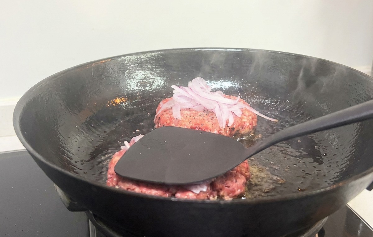 Une spatule noire pressant fermement un steak avec des oignons rouges dans une poêle en fonte chaude.