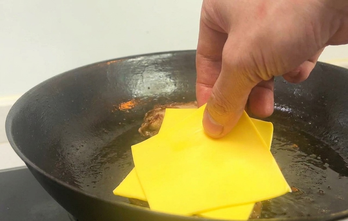 Une main déposant deux tranches carrées de fromage jaune sur les steaks cuits dans une poêle chaude.