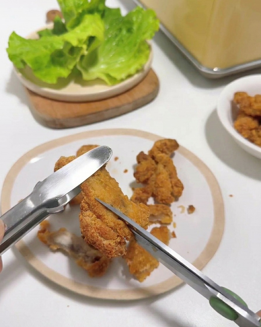 Gros plan de ciseaux de cuisine découpant un morceau de poulet frit chaud tenu par une pince sur une assiette blanche.