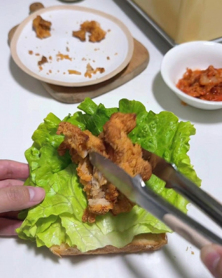 Pince déposant des lamelles de poulet frit sur une couche de laitue sur du pain grillé.