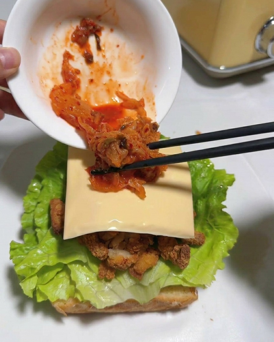 Baguettes transférant du kimchi haché rouge vif dun bol blanc vers une tranche de fromage dans un sandwich.