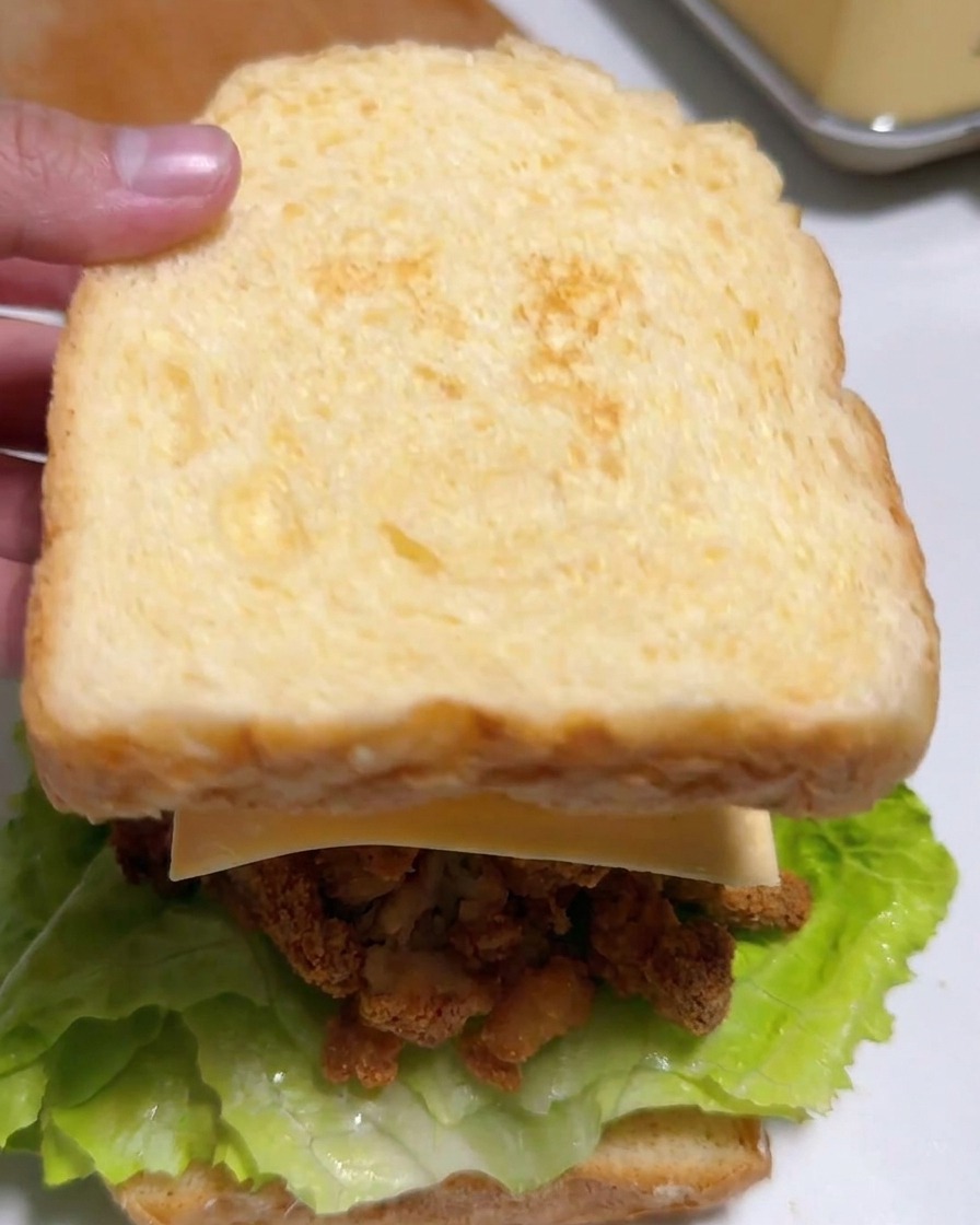 Une main posant la deuxième tranche de pain grillé sur le sandwich garni de poulet, fromage et kimchi.