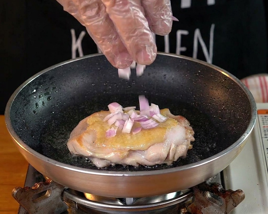 Une main saupoudrant des oignons rouges hachés dans la poêle avec le poulet.