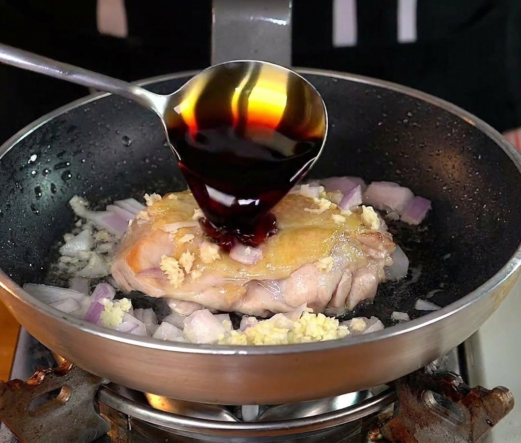 Une cuillère versant de la sauce soja sombre sur le poulet en cours de cuisson.
