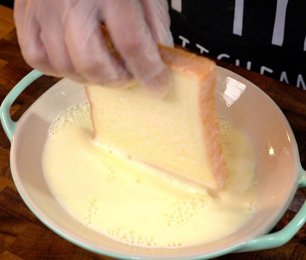 Une main trempant une tranche de pain blanc épaisse dans le mélange dœufs et de lait.