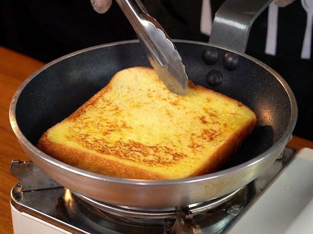 Une tranche de pain perdu bien dorée retournée dans une poêle avec des pinces.