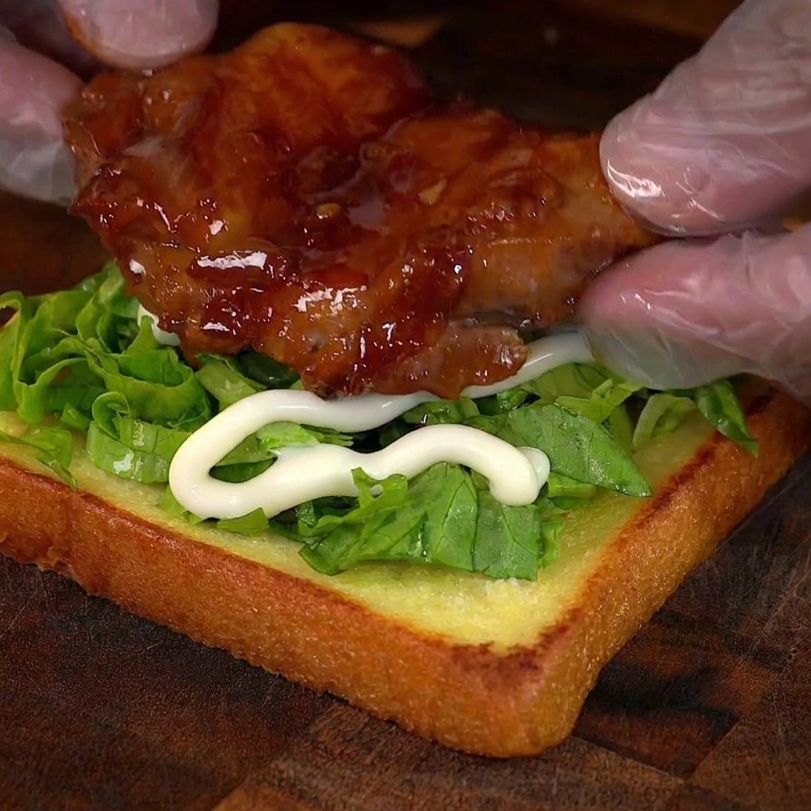 Des mains posant le poulet caramélisé sur une base de pain avec de la laitue.