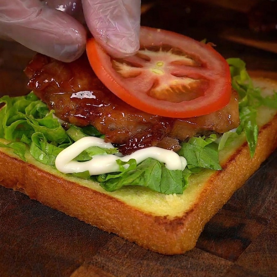 Une main posant une rondelle de tomate fraîche sur le sandwich au poulet.