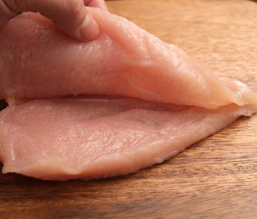 Une main coupant un blanc de poulet cru horizontalement sur une planche en bois.