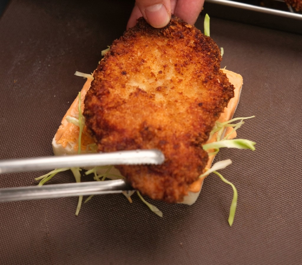 Une pince déposant le poulet frit croustillant sur le pain garni de chou et de sauce.